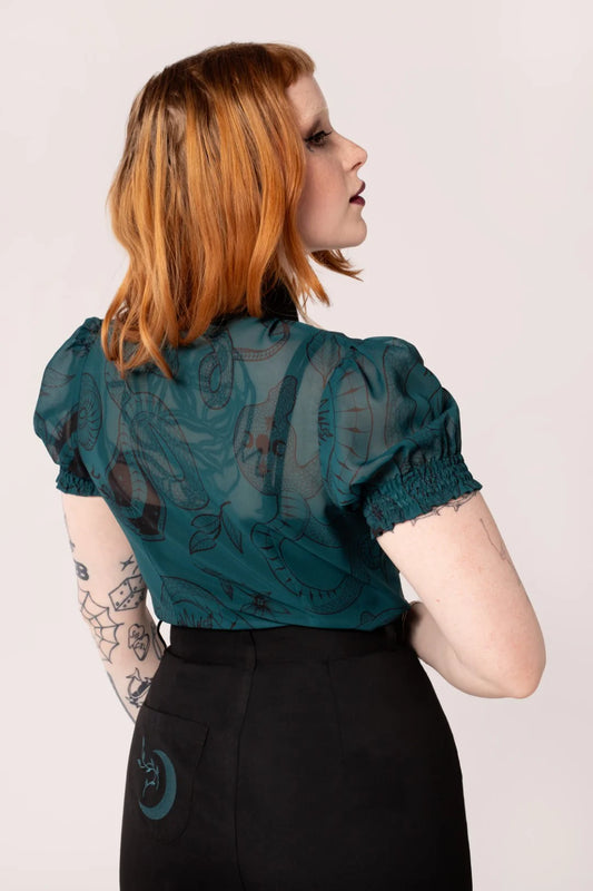 Woman wearing a teal lace top and black pants against a plain background